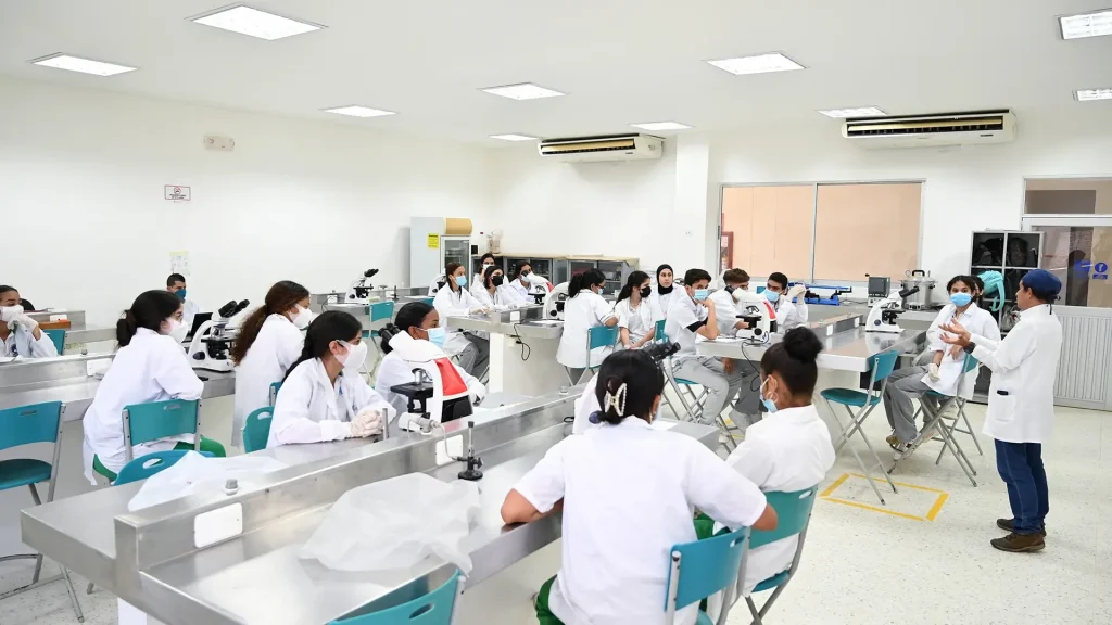 Grupo grande de estudiantes sentados frente a una mesa, con elementos de bioseguridad, en un laboratorio, observando a un docente que está explicando.
