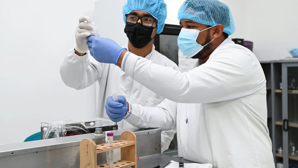 Dos estudiantes en un laboratorio de biología, vestidos con batas y gorros, manejando tubos de ensayo y equipos.