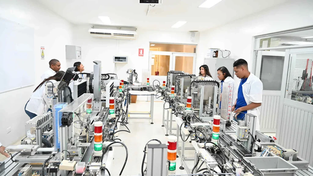 Un grupo de estudiantes en batas blancas trabaja en un laboratorio de ingeniería o robótica, con máquinas y equipos industriales a lo largo de las mesas.
