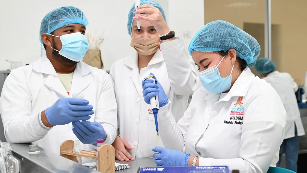 Tres estudiantes en un laboratorio de biología, vestidos con batas y gorros, manejando tubos de ensayo y equipos