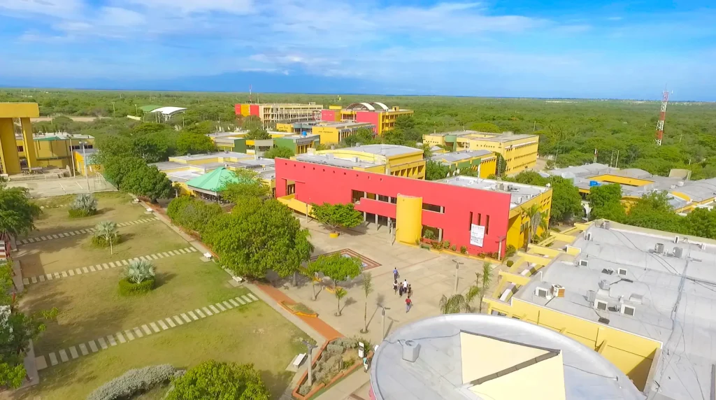 Vista aérea del campus de la Universidad de La Guajira, con edificios amarillos y un edificio rojo central, rodeados de vegetación.