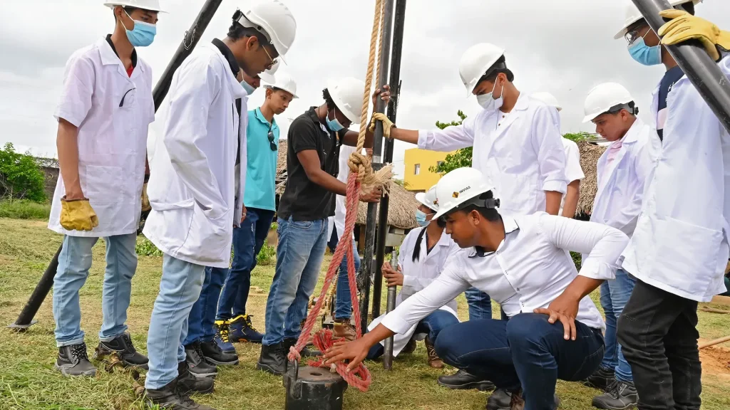 Diez jóvenes estudiantes y un docente, vestidos con batas blancas y cascos de seguridad, se encuentran en las instalaciones de la Universidad de La Guajira realizando prácticas de campo sobre estudios de suelo.