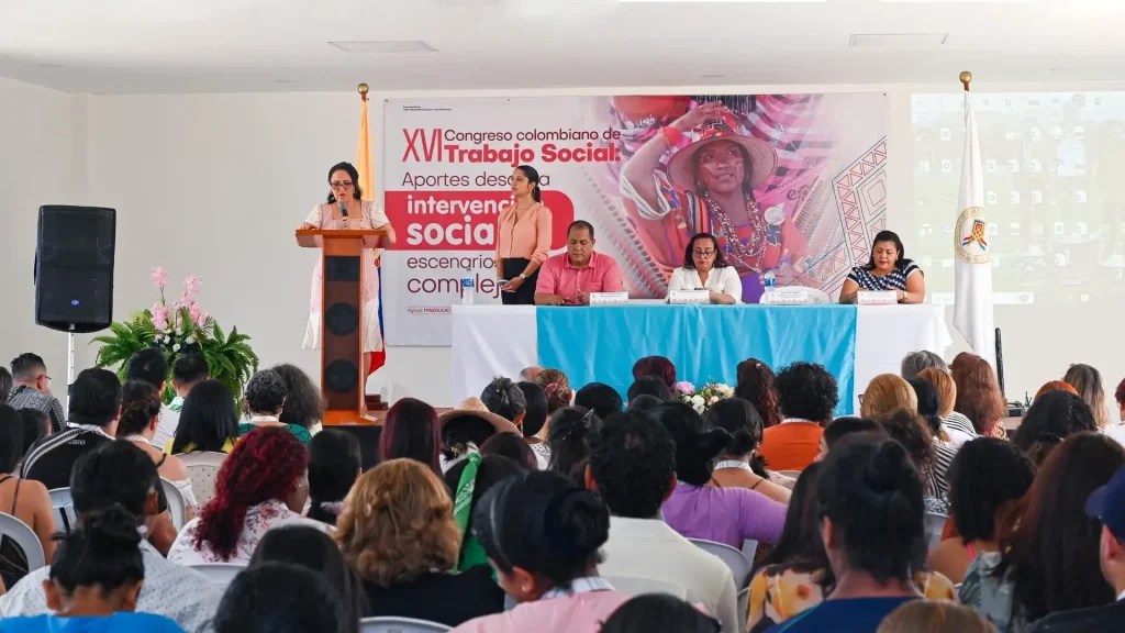 Auditorio lleno de personas concentradas, mientras que está una mujer adulta hablando con un micrófono en el atril. Adicionalmente está una mujer en la tarima de pie con las manos cruzadas y tres personas sentadas en una mesa.