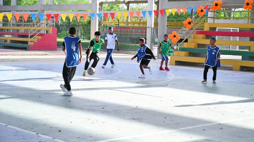 5 niños jugando fútbol en una cancha y un hombre jóven con uniforme está arbitrando el partido.