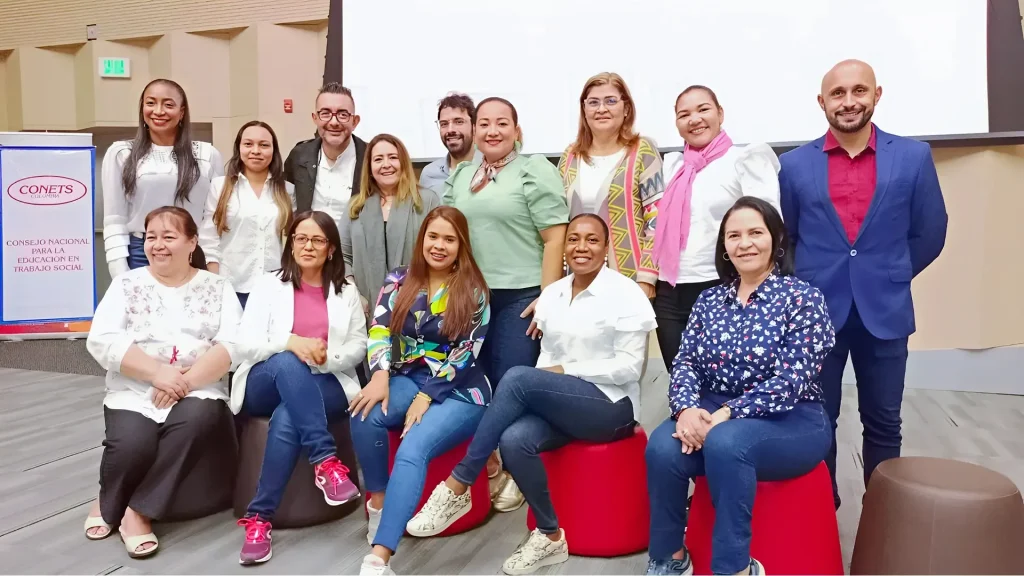Grupo de personas en un auditorio, vestidos de manera formal, 5 de ellas se encuentran sentadas en unos puff.