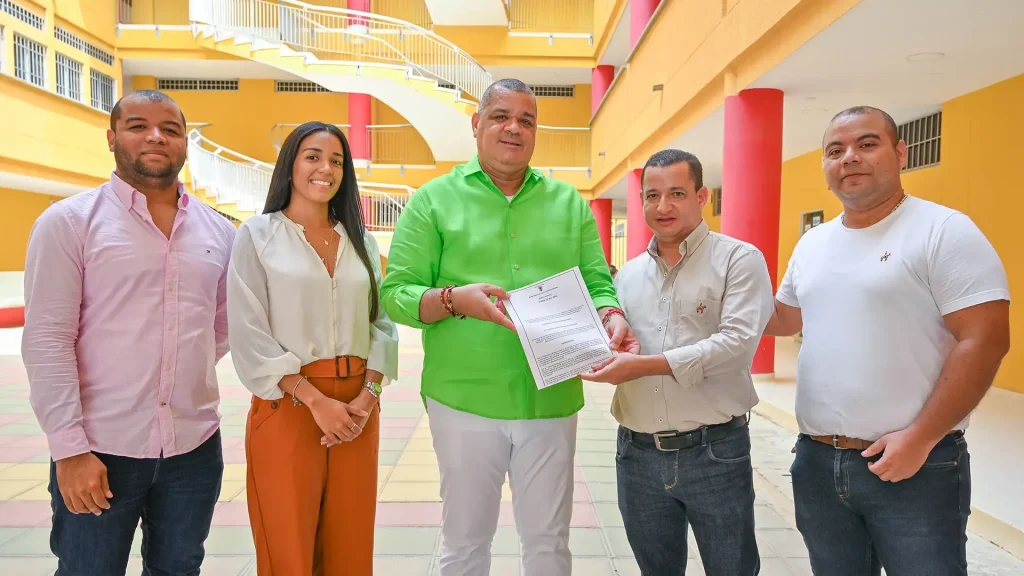 Cuatro hombres y una mujer en un entorno educativo, dos de ellos sostienen un documento.