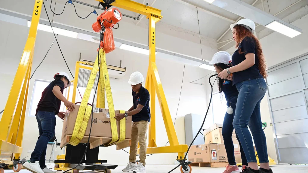 Cuatro estudiantes con cascos de protección, operando un montacargas para mover una caja grande en lo que parece un taller.