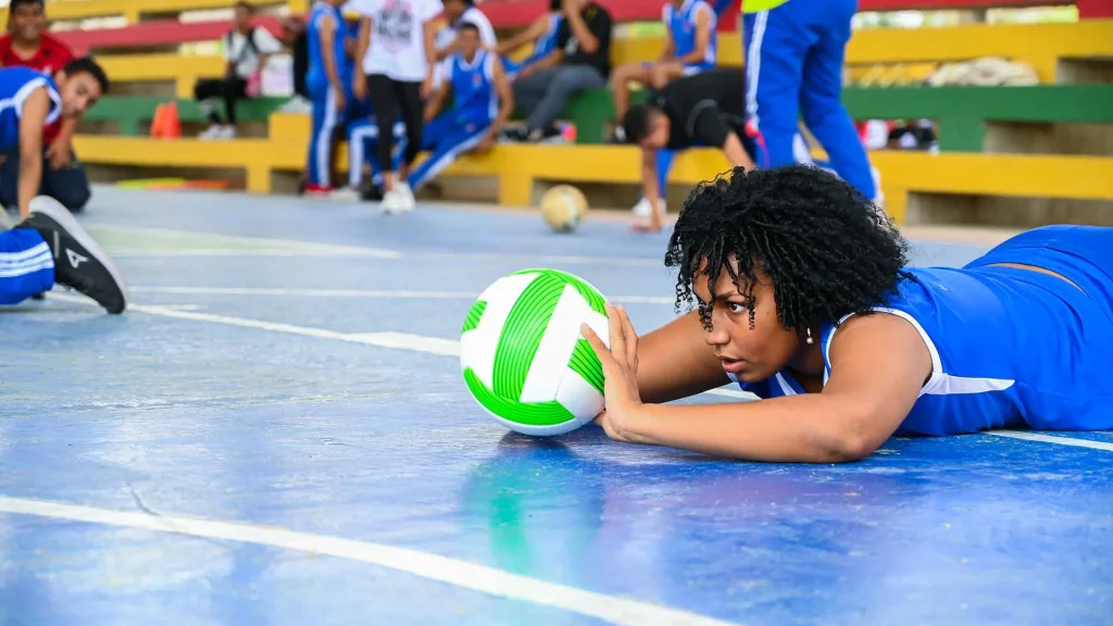 Una mujer joven con uniforme deportivo azul se lanza al suelo para alcanzar un balón de voleibol.