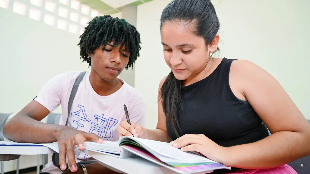 Un hombre y una mujer jóvenes sentados en un salón de clases, mirando un cuaderno y tomando notas juntos.