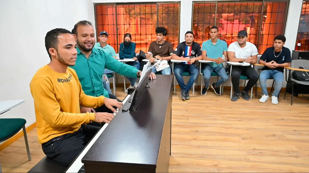 Un instructor de música ayuda a un estudiante a tocar un piano electrónico en un salón de clases con otros estudiantes observando.