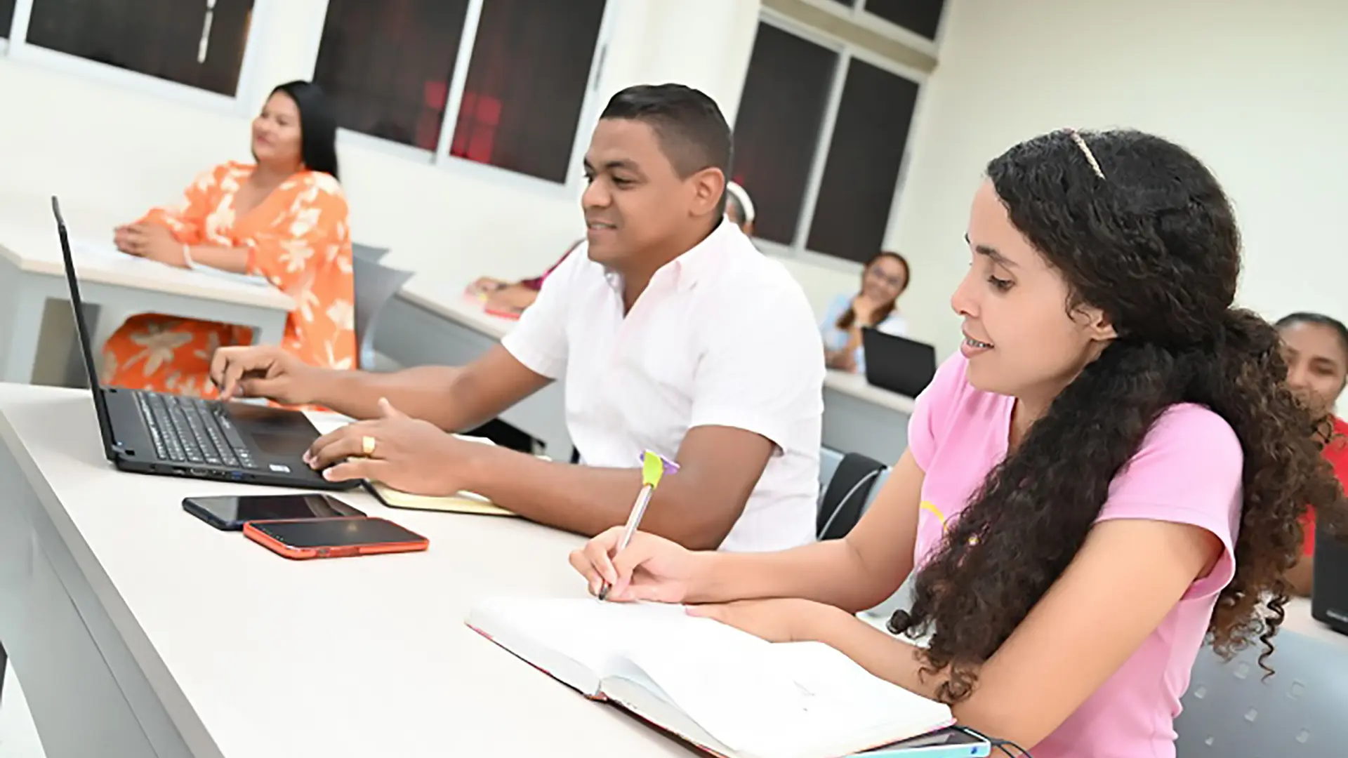 Dos estudiantes, una chica escribiendo en un cuaderno y un joven trabajando en un portátil, sentados juntos en un aula.