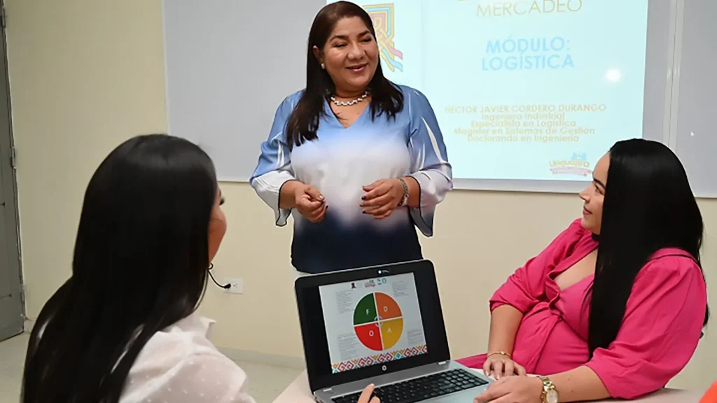 Una profesora dando una clase a dos estudiantes con una laptop en el escritorio.
