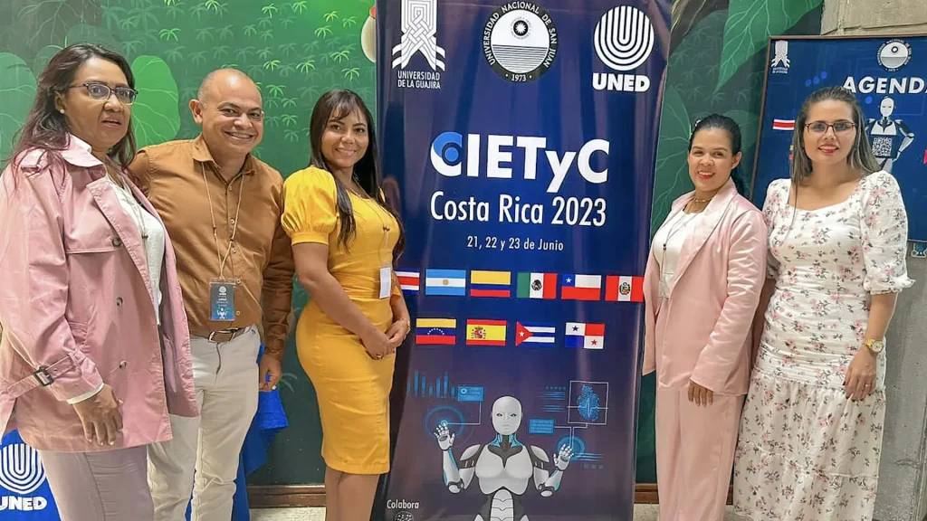 Cuatro mujeres y un hombre adulto posan para una foto junto a un poster de un congreso
