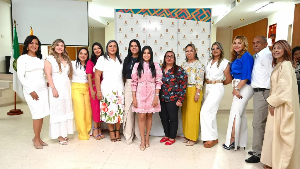 Un grupo de 13 personas en donde la mayoría son mujeres y solo un hombre posan para una foto, en un lugar cerrado, en el fondo de la imagen se alcanza a visualizar una bandera color verde con blanco que representaría el símbolo oficial del departamento de la Guajira.