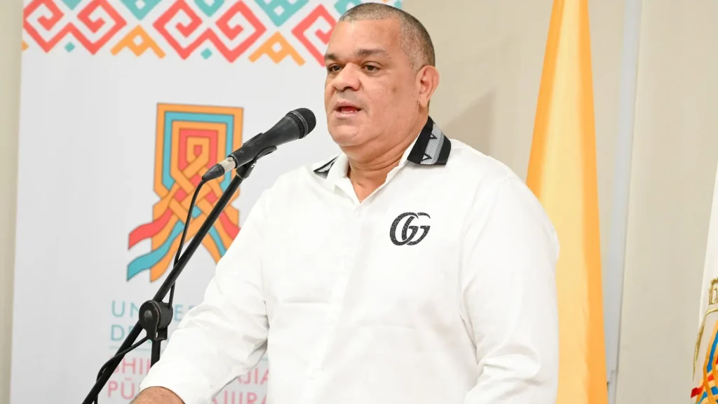 Un hombre vestido con camisa blanca formal, de pie frente a un micrófono, aparentemente dictando una charla, con una bandera atrás color naranja y del lado izquierdo un pendón con el tejido y logo institucional de la Universidad de la Guajira. 