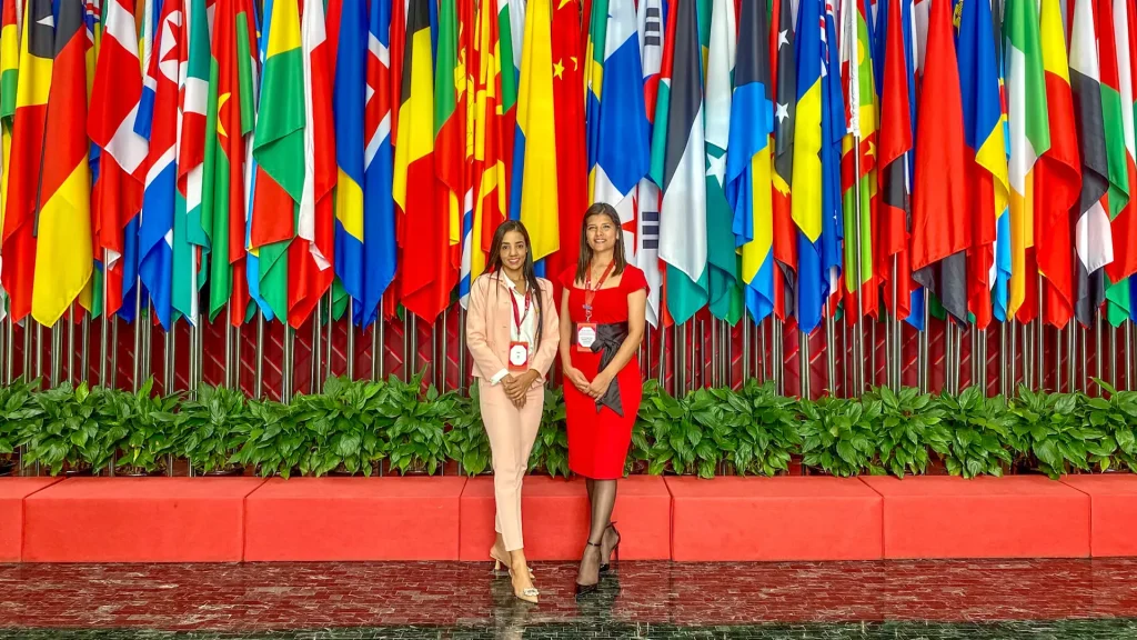 Dos mujeres adultas posan para una foto delante de las banderas de los países, al interior de un auditorio.