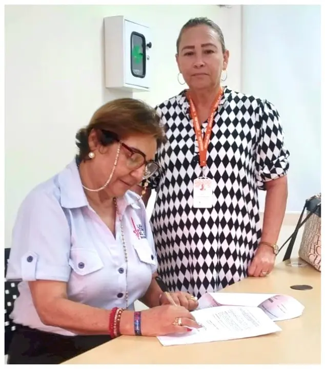 Dos mujeres una de pie mirando la cámara y otra sentada firmando unos papeles mientras se apoya de una mesa