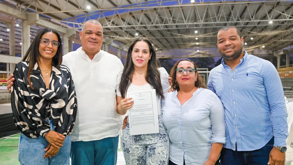 Dos hombres y tres mujeres sonriendo en un coliseo de noche. La mujer del centro se encuentra sosteniendo un certificado.