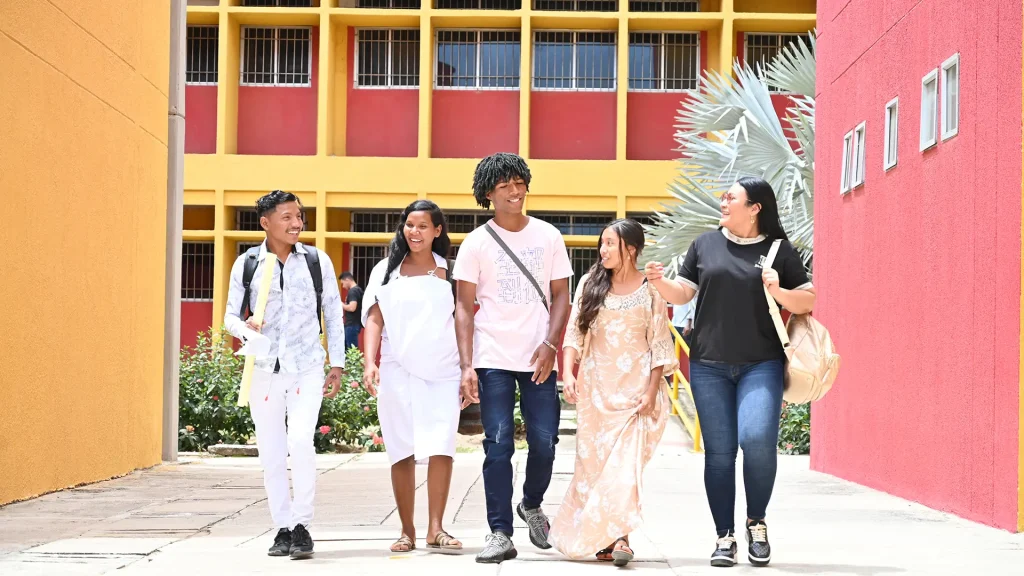 Un grupo de cinco estudiantes sonrientes camina al aire libre entre edificios de colores brillantes, uno amarillo y otro rojo.