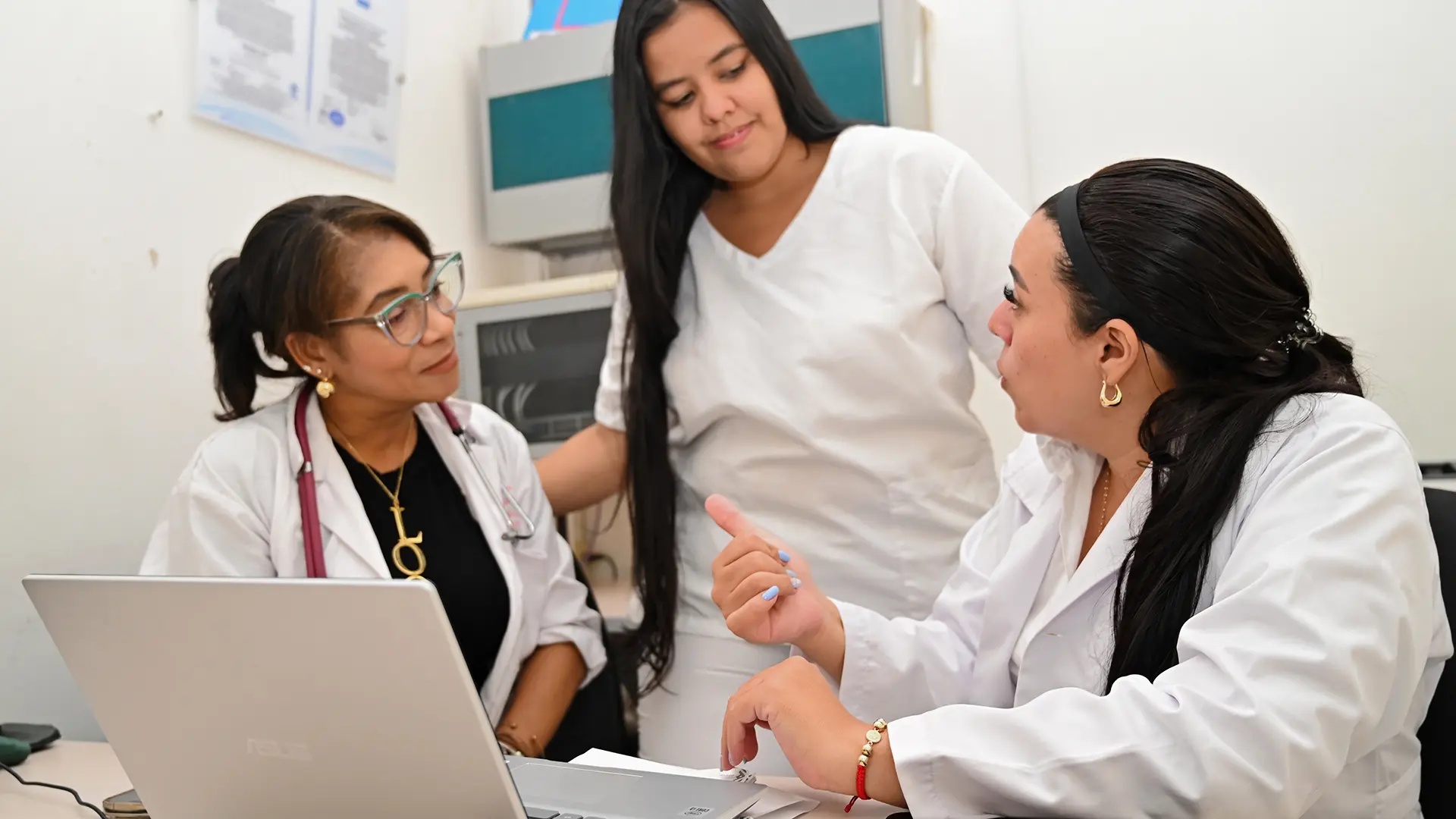 Tres mujeres con vestimenta alusiva a actividades de la salud. Están en un consultorio hablando entre ellas y al frente tienen un computador abierto.