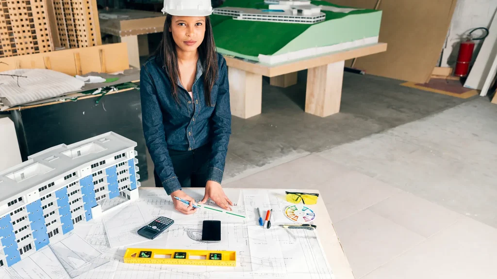 Mujer joven, con camisa de jean manga larga. Tiene un casco de protección blanco y tiene sus manos colocadas sobre unas maquetas de arquitectura.