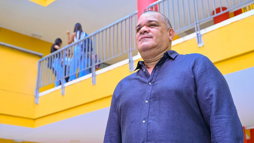 El rector de la Universidad de La Guajira, Carlos Robles Julio, de pie en un bloque universitario. Viste una camisa azul y mira hacia un lado, con un fondo de paredes amarillas y un balcón donde se ven figuras borrosas de estudiantes.