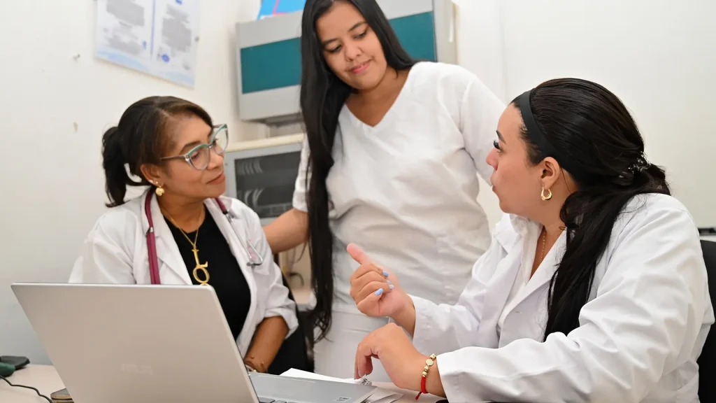 Tres mujeres, probablemente doctoras, están en un consultorio. Dos de ellas conversan mientras la tercera mira una computadora portátil.