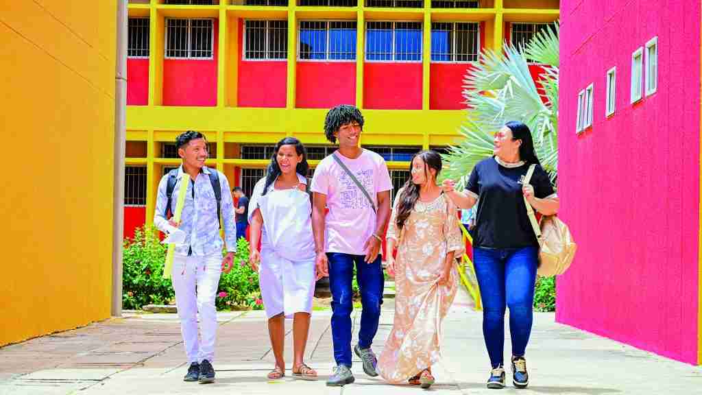 Un grupo de cinco jóvenes estudiantes camina y sonríe por un campus universitario, con edificios de colores brillantes como fondo.