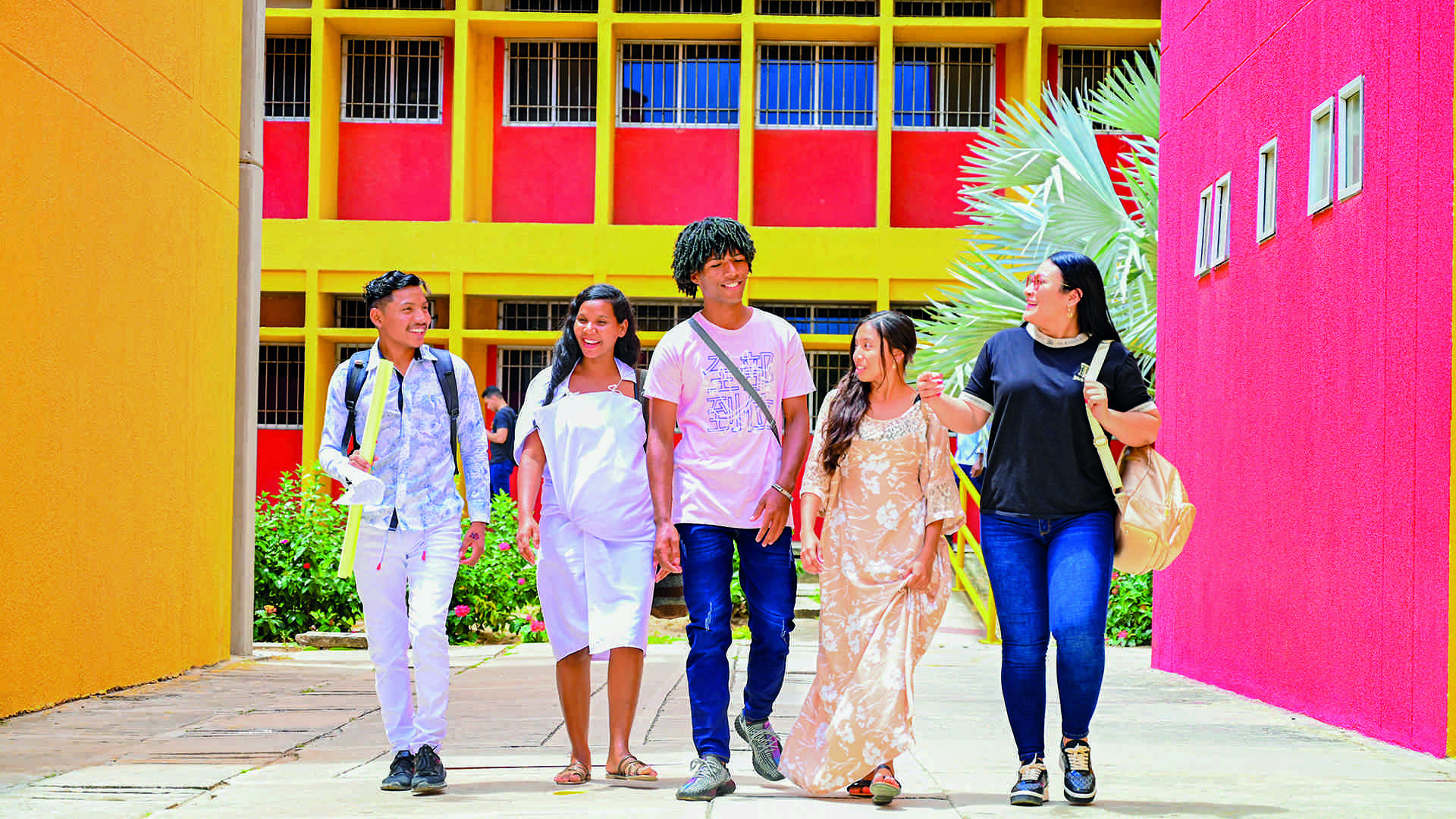 Un grupo de cinco jóvenes estudiantes camina y sonríe por un campus universitario, con edificios de colores brillantes como fondo.