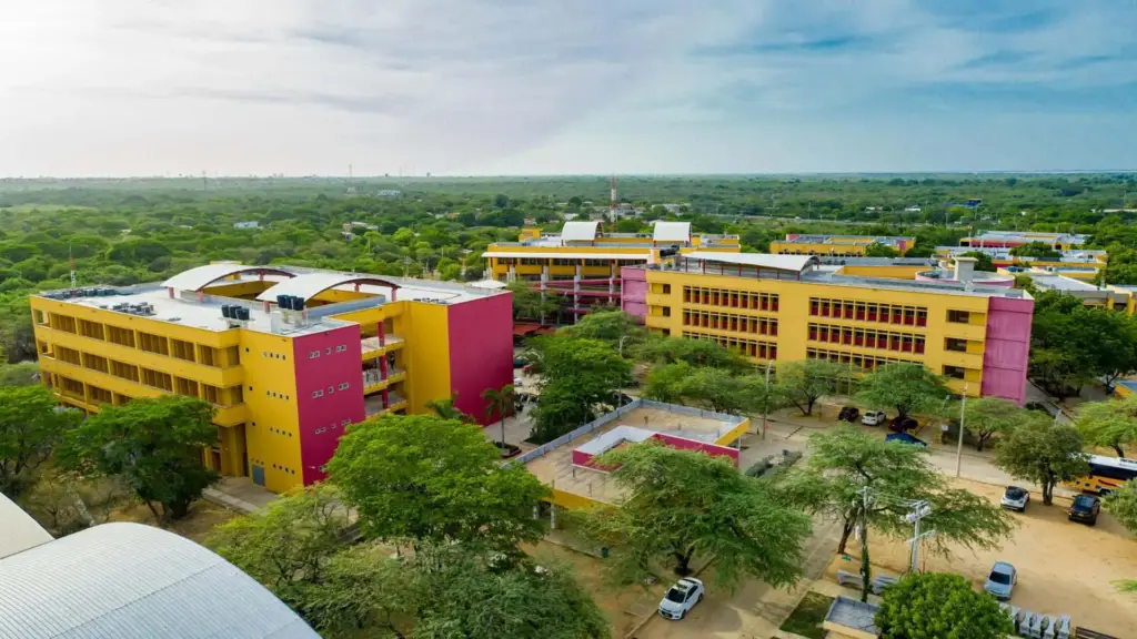 Vista aérea del campus de la Universidad de La Guajira, mostrando varios edificios modernos de colores rojo y amarillo, rodeados de vegetación.