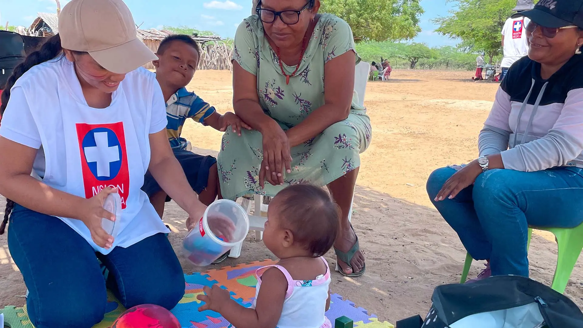 Una mujer joven interactúa con un bebé, mientras otra mujer mayor y un niño pequeño observan. La escena tiene lugar al aire libre bajo una estructura rústica de sombra en un entorno rural desértico