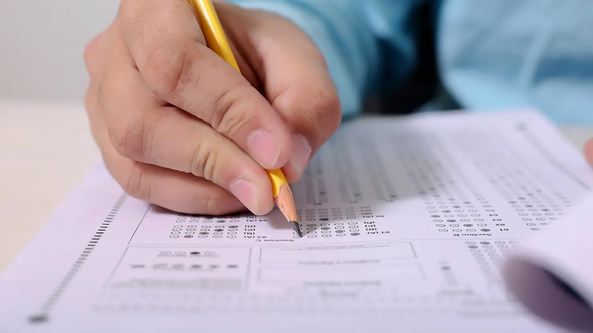 Persona llenando un examen tipo prueba estandarizada con un lápiz. Se observan círculos de respuestas de opción múltiple en la hoja de respuestas, con algunas secciones ya completadas. La persona utiliza un lápiz amarillo para marcar las opciones.