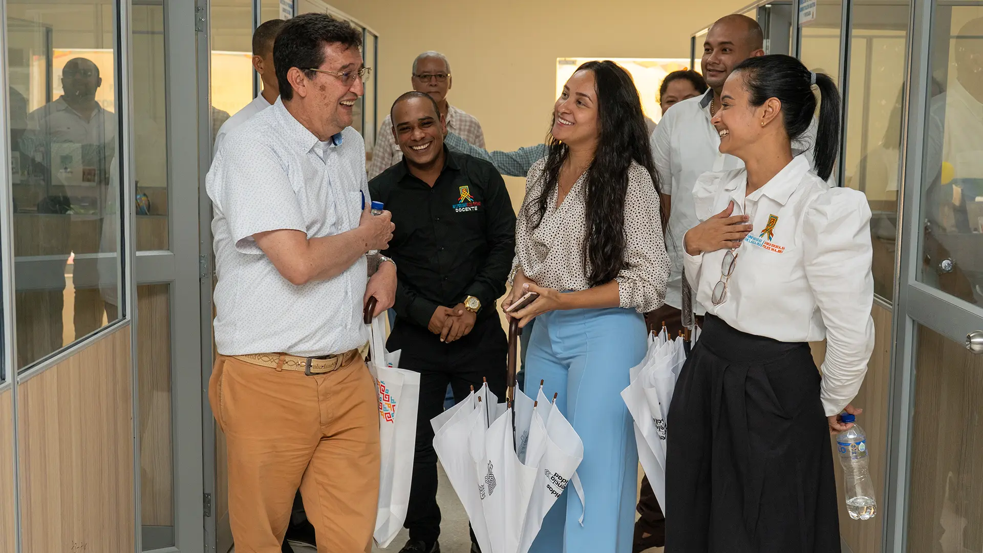 Un hombre de camisa negra con un logotipo bordado da una presentación frente a una pantalla. Usa un reloj dorado y gesticula con las manos mientras explica, en un ambiente de aula o sala de conferencias.