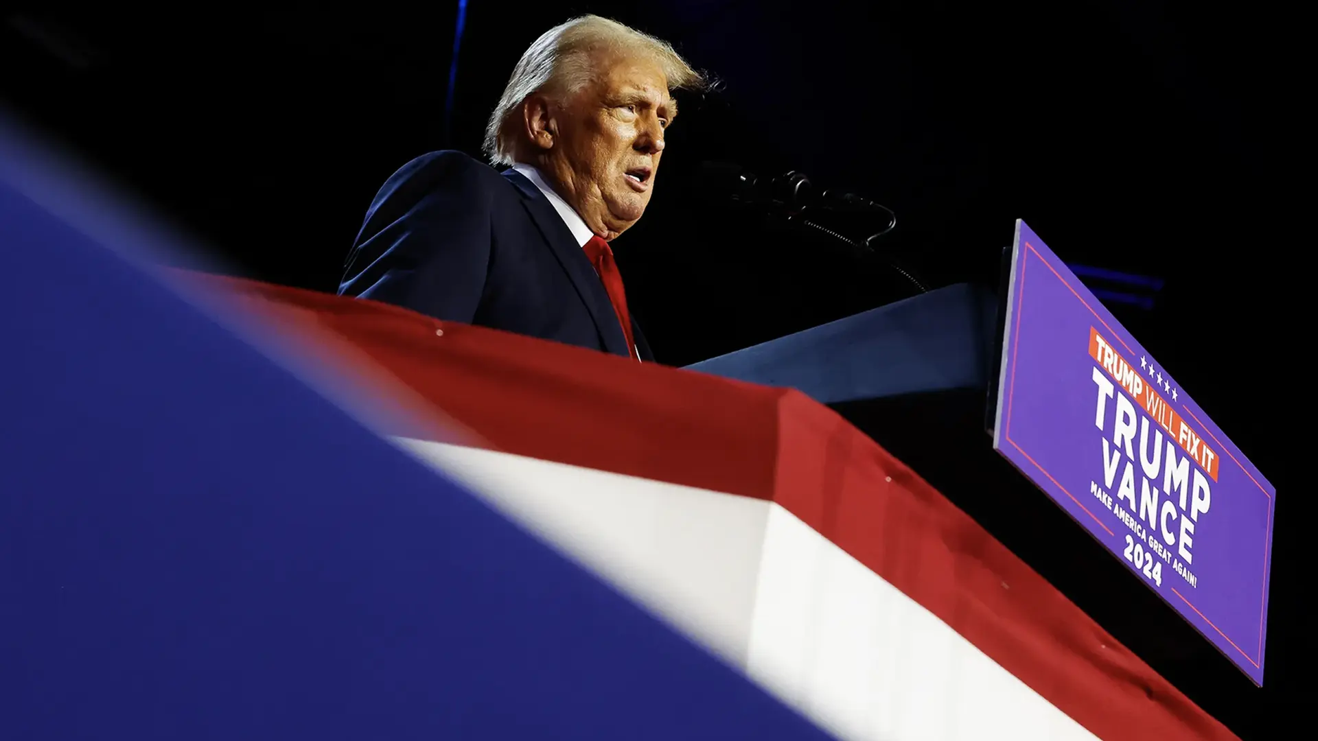 Donald Trump en un podio decorado con una bandera de EE. UU., dando un discurso. En la esquina derecha, un cartel dice "Trump Will Fix It - Trump Vance - Make America Great Again! 2024", indicando una campaña política en curso. La persona está vestida con un traje oscuro y corbata roja, iluminada por luces que resaltan su perfil contra un fondo oscuro