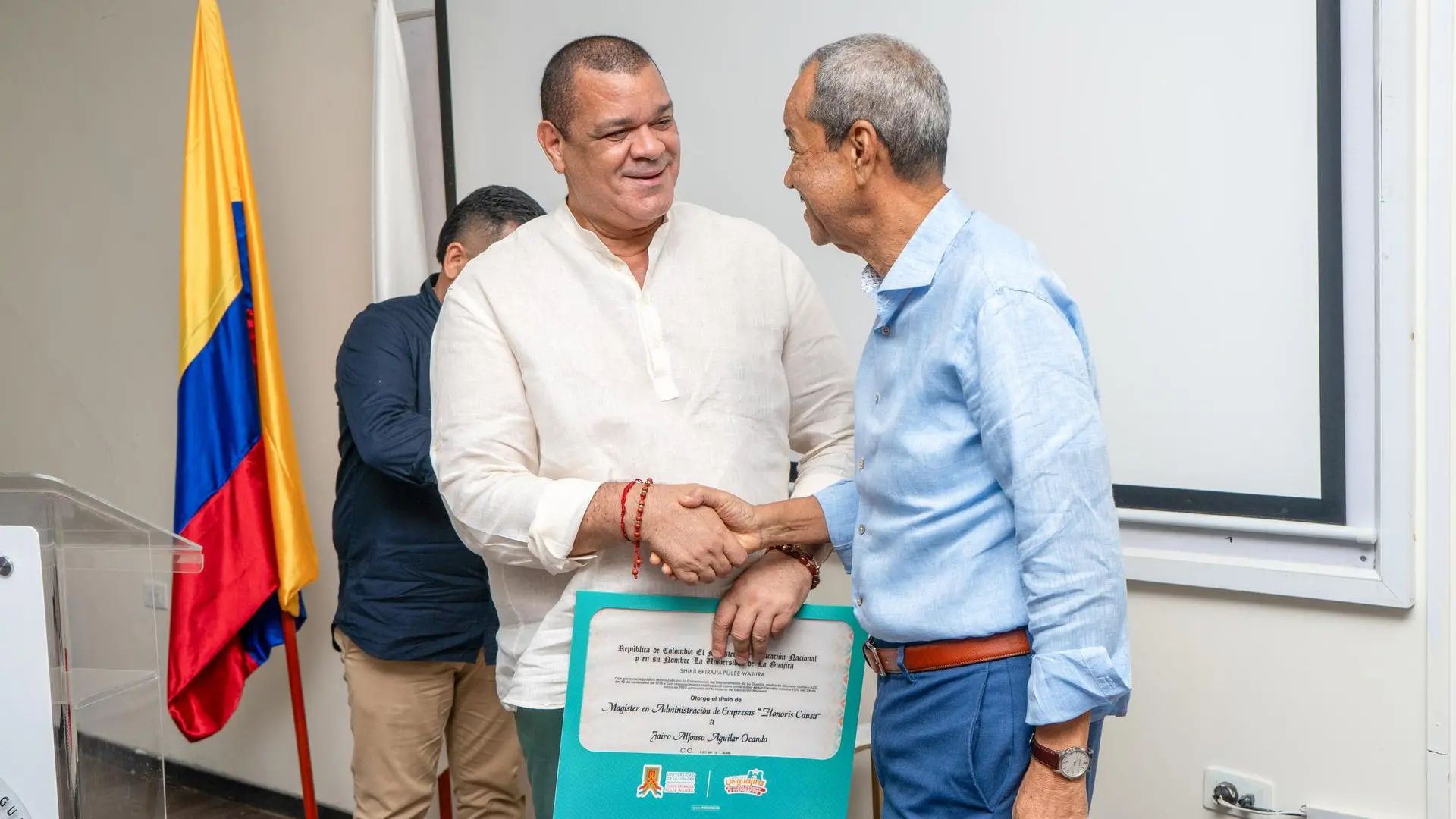 Dos hombres estrechándose las manos en un evento formal. Uno de ellos sostiene un diploma o reconocimiento. De fondo, se puede ver la bandera de Colombia y una pantalla blanca.