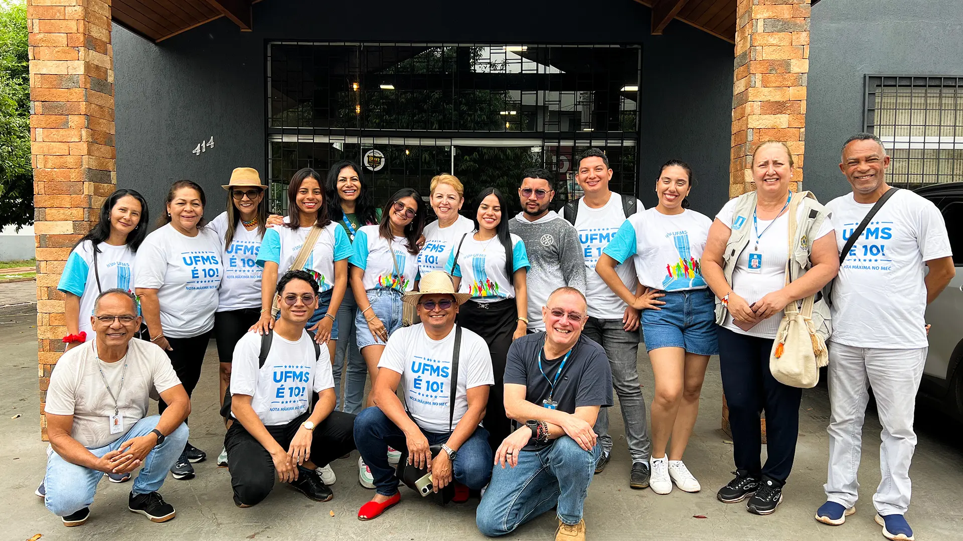 Un grupo diverso de personas, en su mayoría usando camisetas blancas con el logo "UFMS É 10!", posa frente a un edificio de ladrillos y pintura oscura. Están sonriendo y parecen estar participando en un evento o actividad comunitaria.