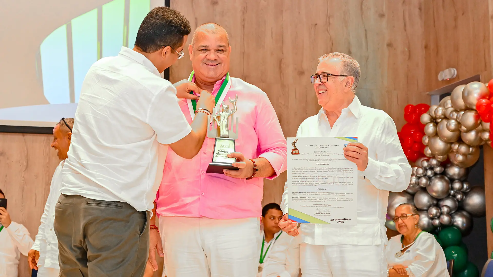 Un hombre con camisa rosa está recibiendo una medalla verde alrededor del cuello durante una ceremonia de premiación. Sostiene un trofeo plateado y un certificado en la mano. A su lado, otro hombre de camisa blanca sonríe mientras sujeta el certificado. En el fondo hay decoración de globos metálicos y personas observando el momento.