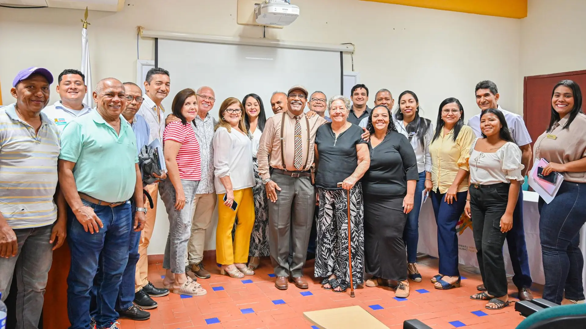 Un grupo diverso de personas posando juntas en un aula. La mayoría sonríe y se encuentran en un ambiente de aprendizaje o reunión. En el fondo, se observa una pantalla de proyección y banderas.