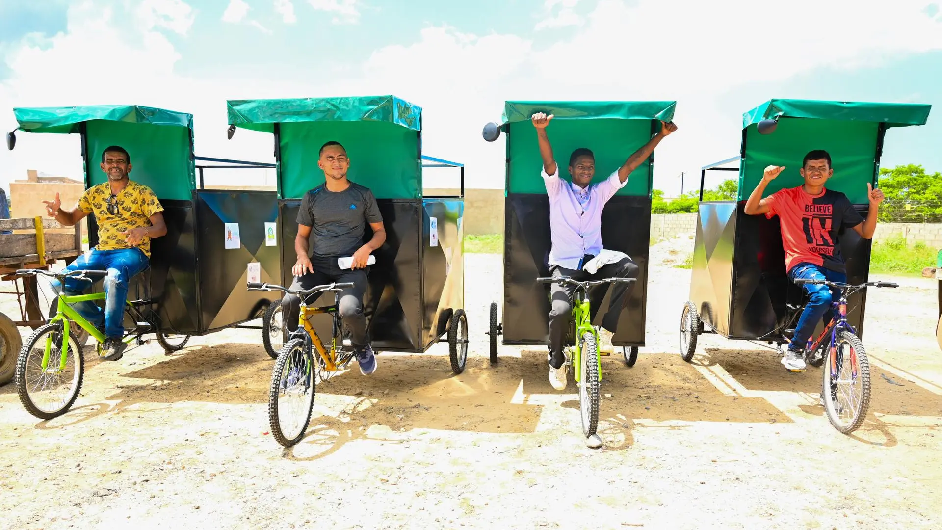 Cuatro hombres jóvenes posan sonrientes sobre triciclos de carga con techo verde, alineados en fila sobre un terreno descubierto en un día soleado.