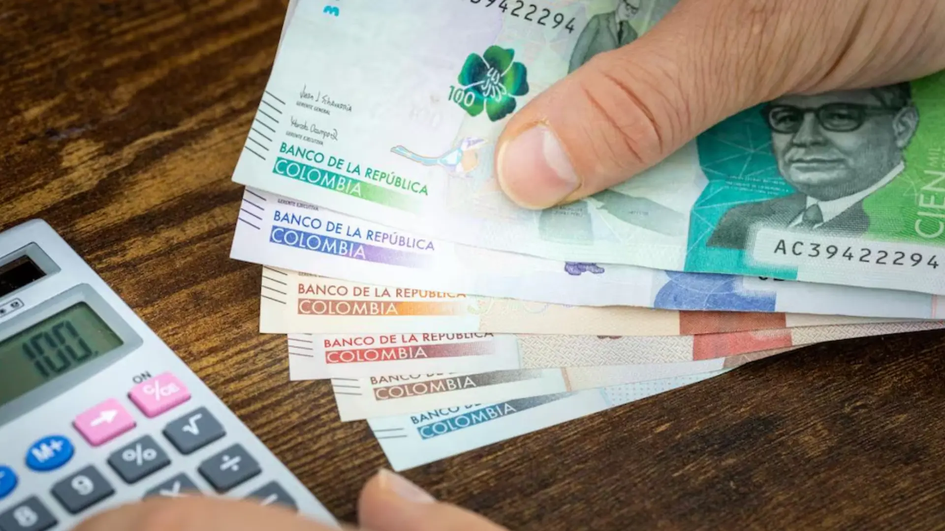 Mano sosteniendo varios billetes colombianos de distintas denominaciones del Banco de la República, colocados en abanico sobre una mesa de madera. A un lado se observa una calculadora, sugiriendo un contexto de cálculo o finanzas personales.