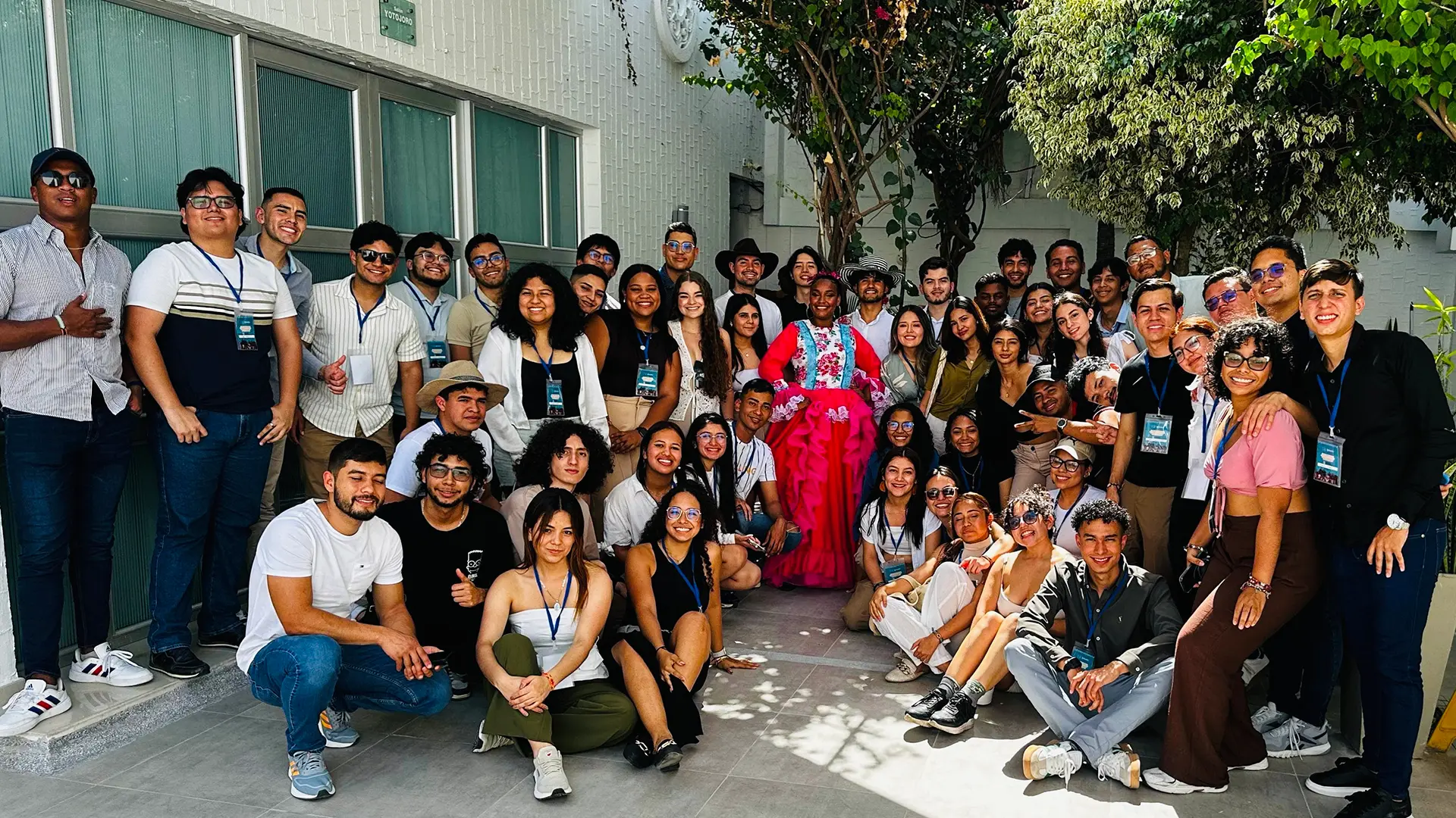 Un numeroso grupo de personas posa sonriente en un patio con vegetación y una pared blanca de fondo. La mayoría lleva gafetes colgados al cuello, lo que sugiere que forman parte de un evento o conferencia. En el centro destaca una mujer con un llamativo vestido rosa adornado con flores, lo que añade un toque festivo y cultural a la escena. La atmósfera es alegre y relajada, con personas de diversas edades y estilos disfrutando del momento.