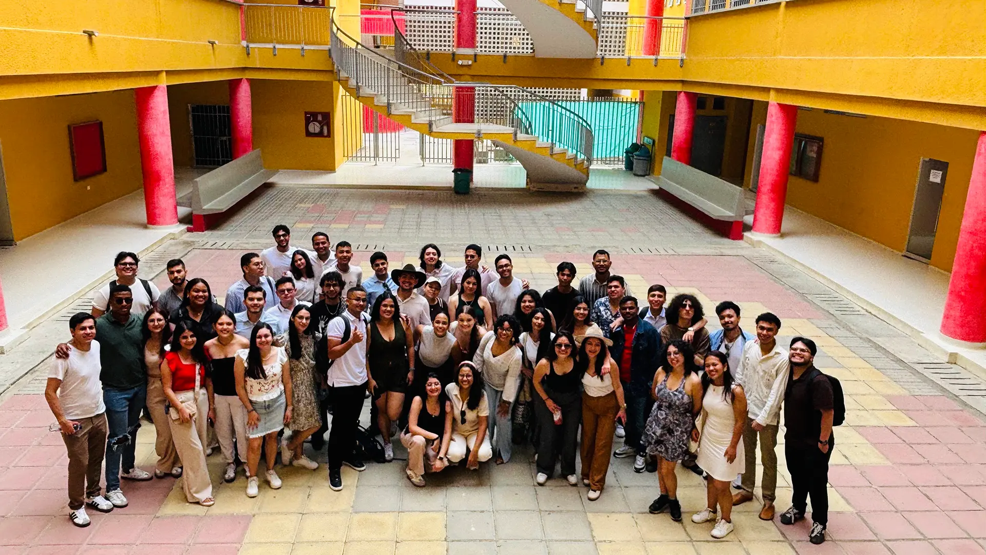 Un grupo de jóvenes posa en un amplio patio interior de un edificio con paredes amarillas y columnas rojas. Al fondo, una escalera con barandales metálicos crea una estructura llamativa. La mayoría de los presentes viste ropa casual y sonríe, reflejando un ambiente de compañerismo y entusiasmo. La disposición del grupo y la arquitectura del lugar aportan profundidad y dinamismo a la escena.