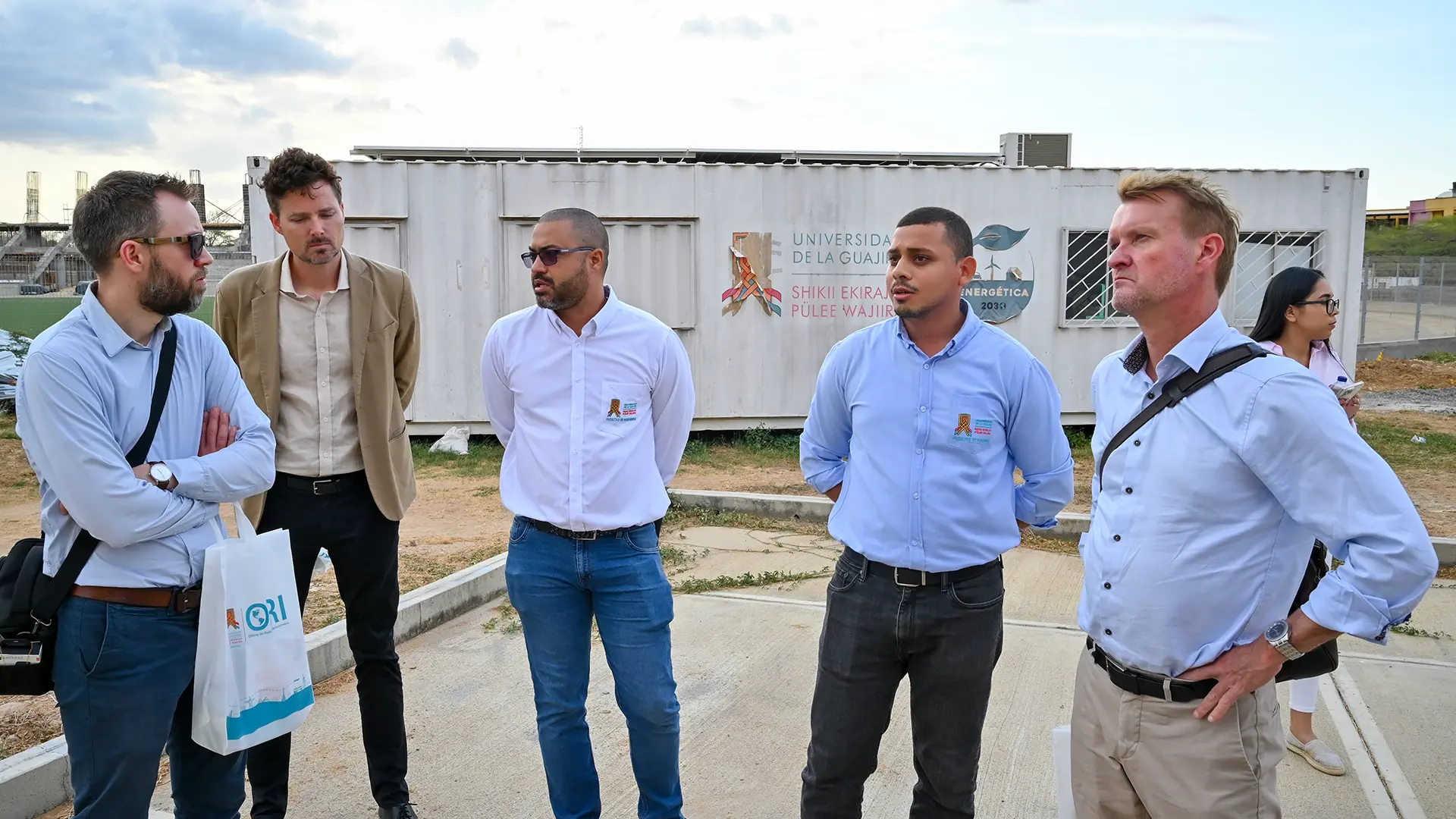 Grupo de profesionales reunidos en exteriores durante una visita institucional a la Universidad de La Guajira, con un contenedor al fondo que forma parte de un proyecto académico o de desarrollo. La interacción refleja un intercambio de ideas en el marco de una colaboración interinstitucional.