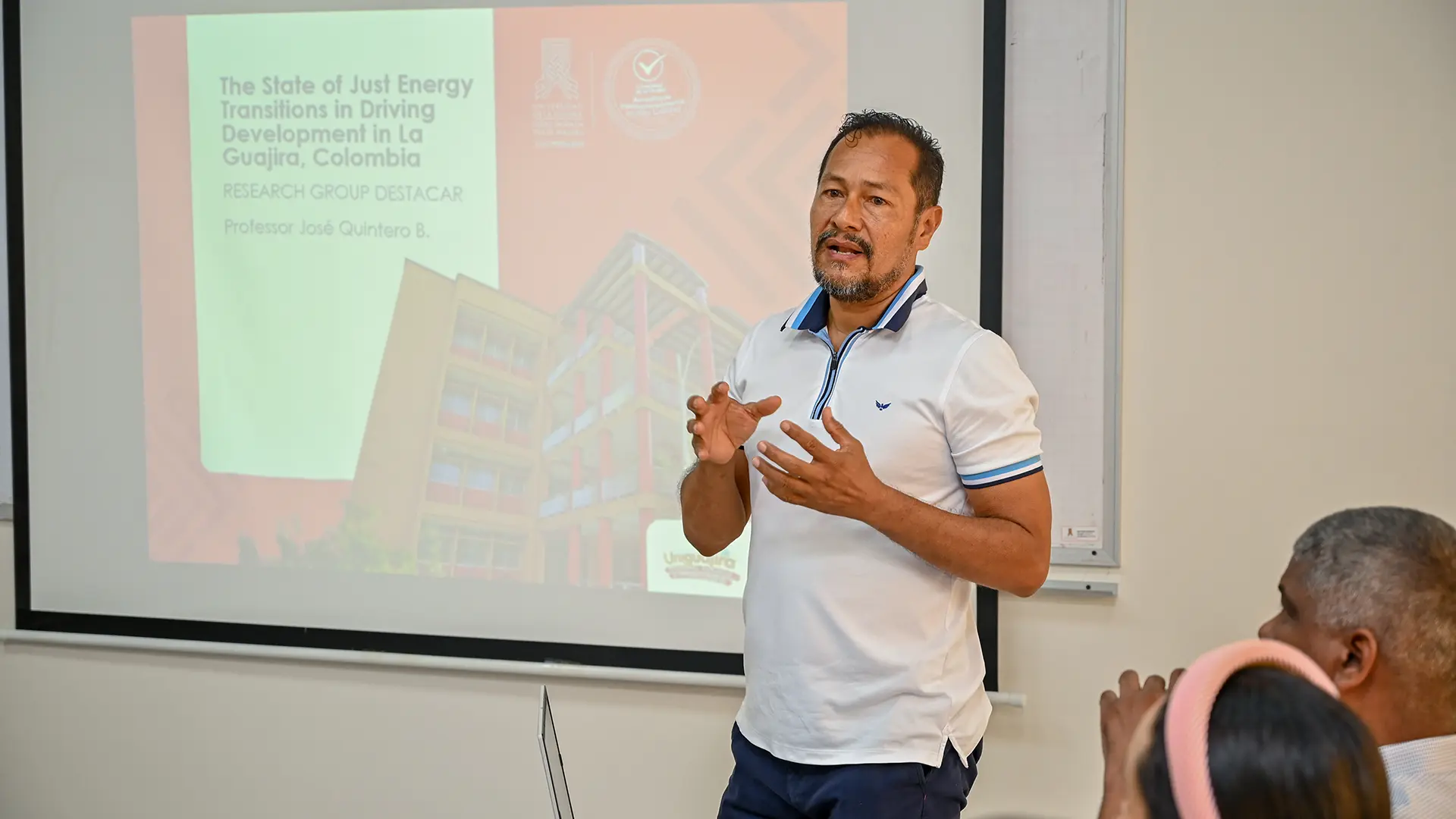 El profesor José Quintero Baldovino presenta los avances del grupo de investigación DESTACAR sobre las transiciones energéticas justas en La Guajira, Colombia, durante una charla en aula. Al fondo se proyecta una diapositiva con el título de su presentación, mientras los asistentes escuchan con atención.