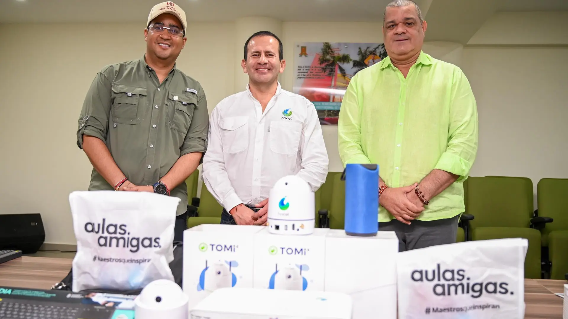 Tres hombres posan sonrientes detrás de una mesa con equipos tecnológicos y bolsas de "Aulas Amigas". La mesa exhibe proyectores, cámaras y dispositivos de aprendizaje. Uno de los hombres viste una camisa con el logo de Hocol.