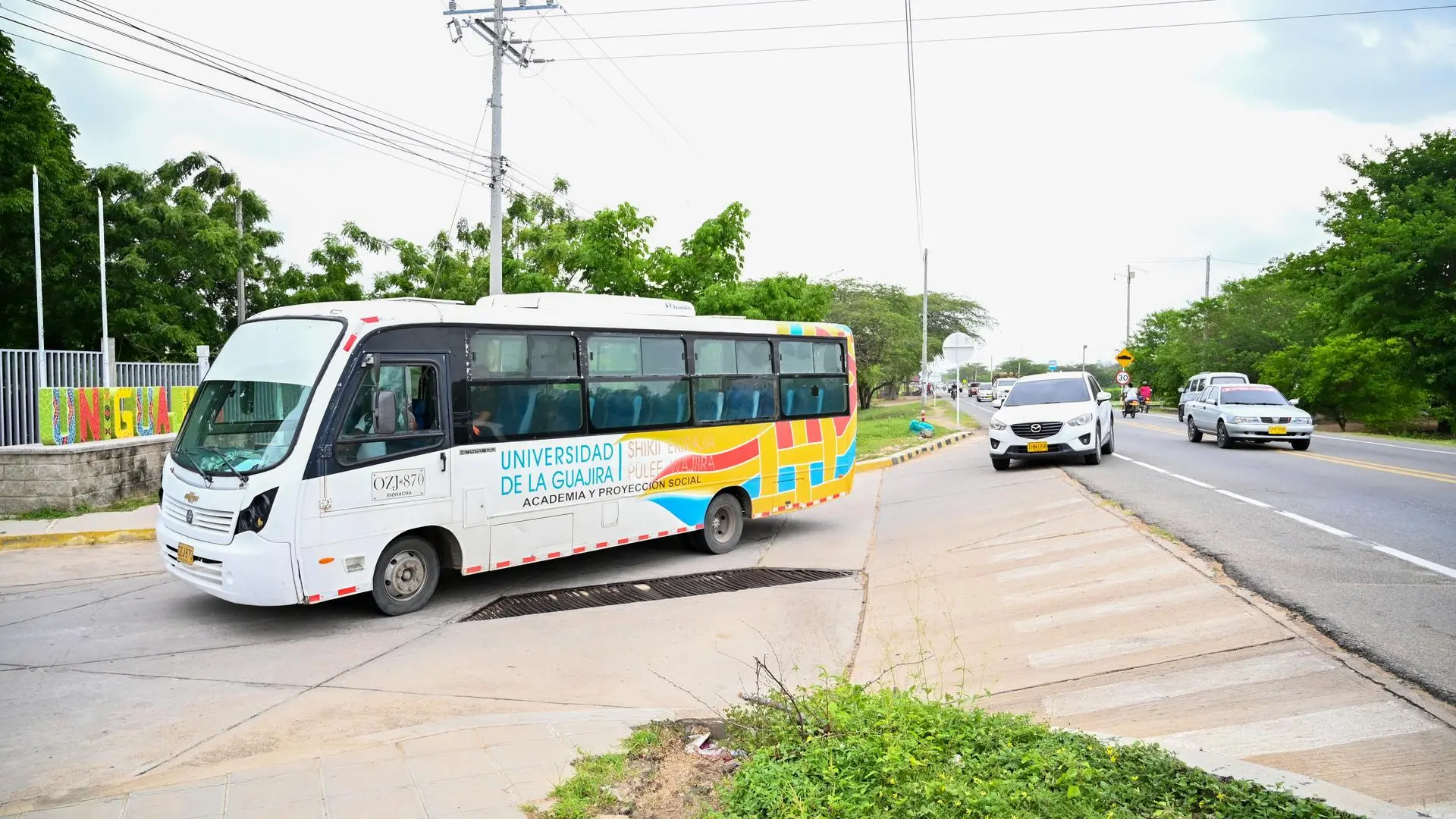 Bus institucional de la Universidad de La Guajira saliendo del campus hacia la vía principal, con árboles al fondo y varios vehículos circulando por la carretera.