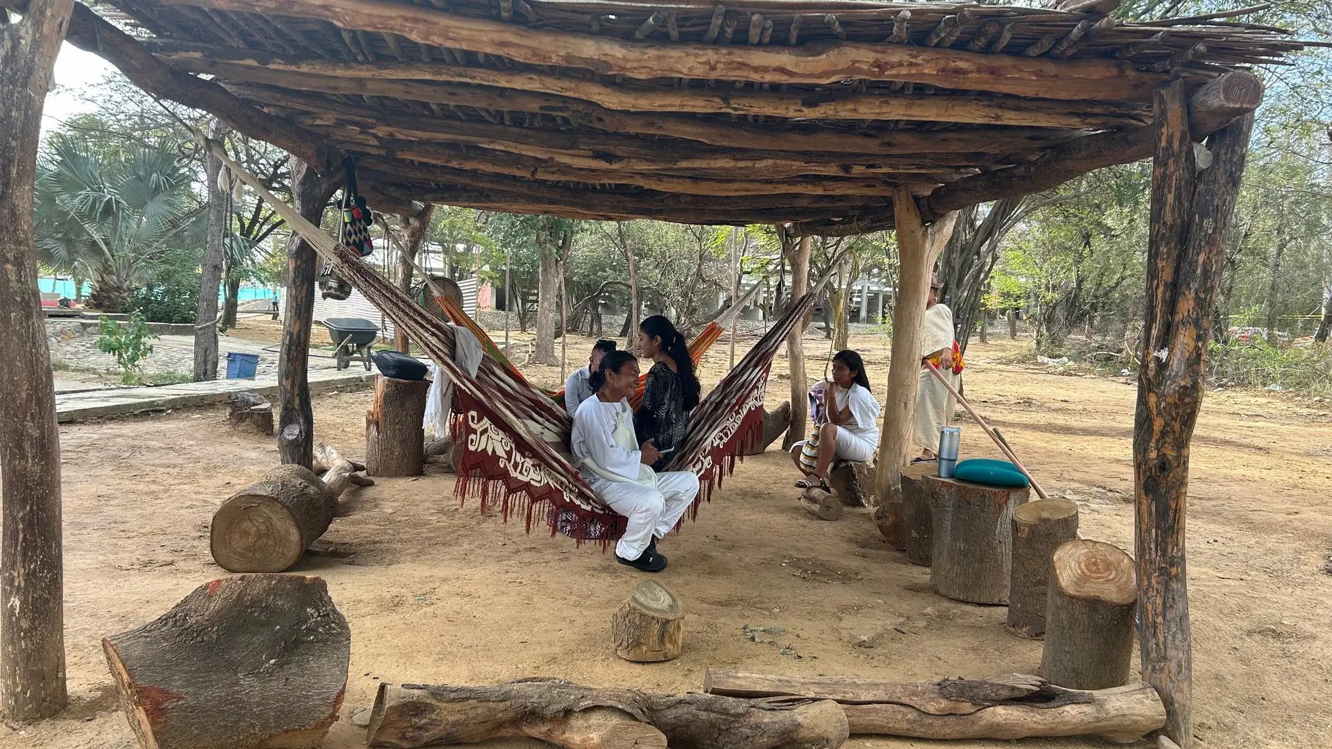 Personas indígenas vestidas con ropa tradicional descansan bajo un techo de madera rústico en un entorno natural. Están sentadas en hamacas tejidas y troncos de árbol usados como bancos. El lugar está rodeado de árboles y tierra seca, con un ambiente tranquilo y comunitario.