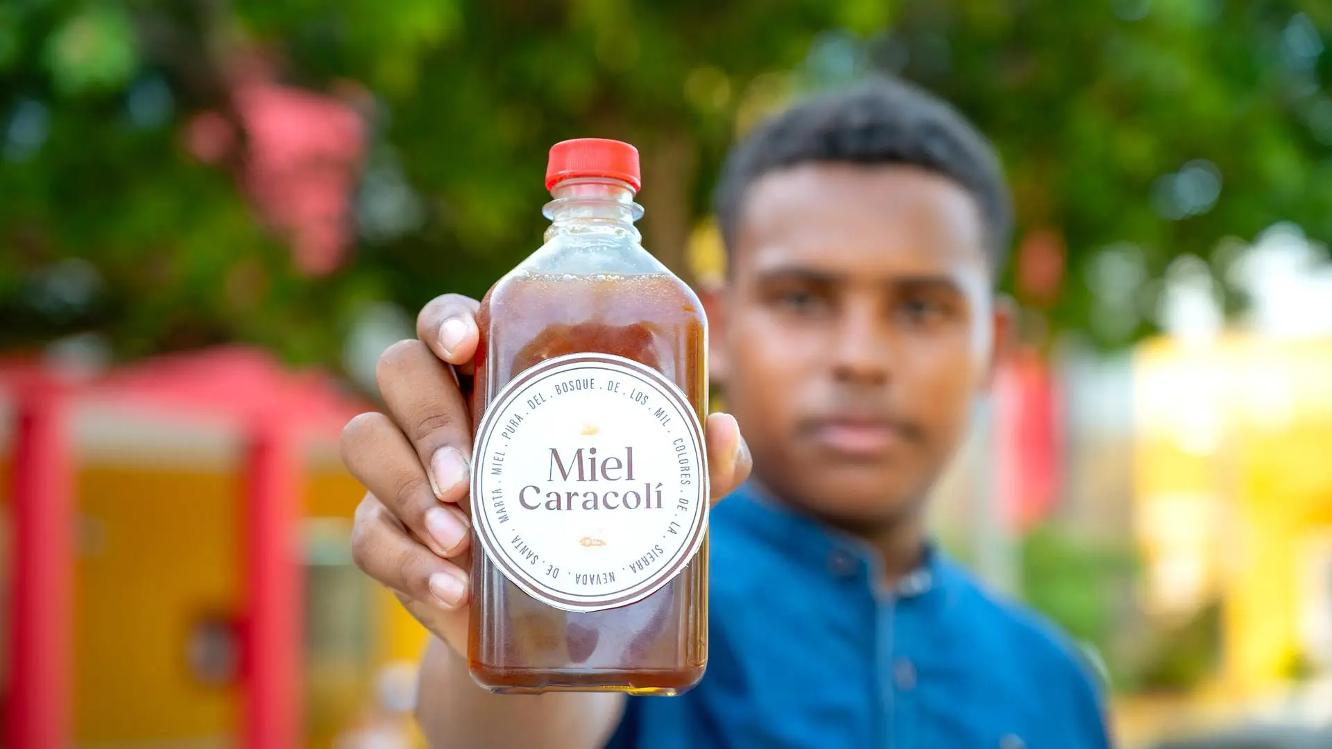 Joven sostiene en primer plano una botella de miel etiquetada como “Miel Caracolí”. La botella tiene tapa roja y contiene miel ámbar. Al fondo, el joven aparece ligeramente desenfocado, con un entorno colorido y árboles, destacando la autenticidad del producto artesanal.
