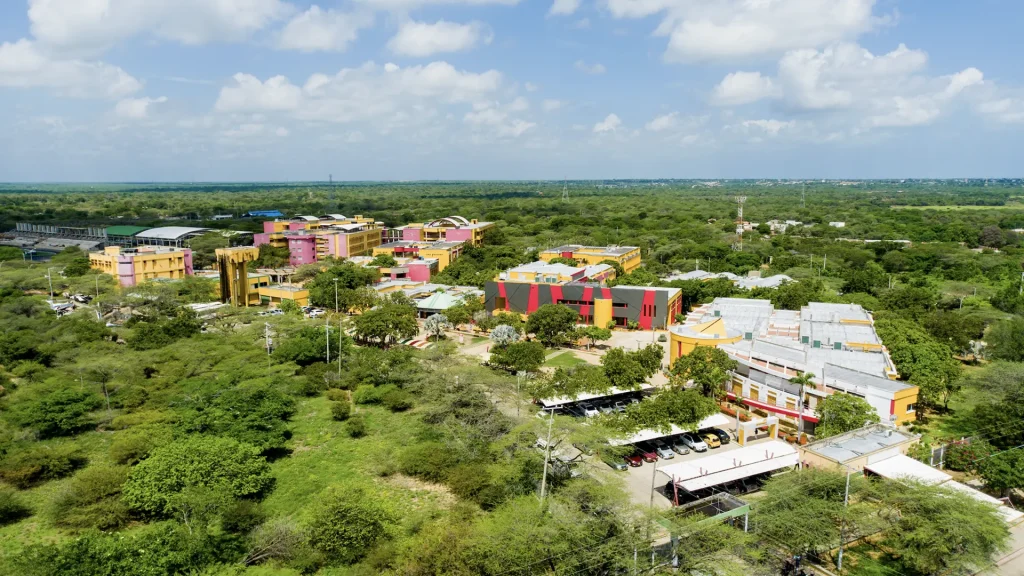 Vista aérea de un campus universitario rodeado de vegetación, con edificios de colores vivos como amarillo, rosa, rojo y negro, distribuidos entre zonas verdes y árboles, bajo un cielo parcialmente nublado.