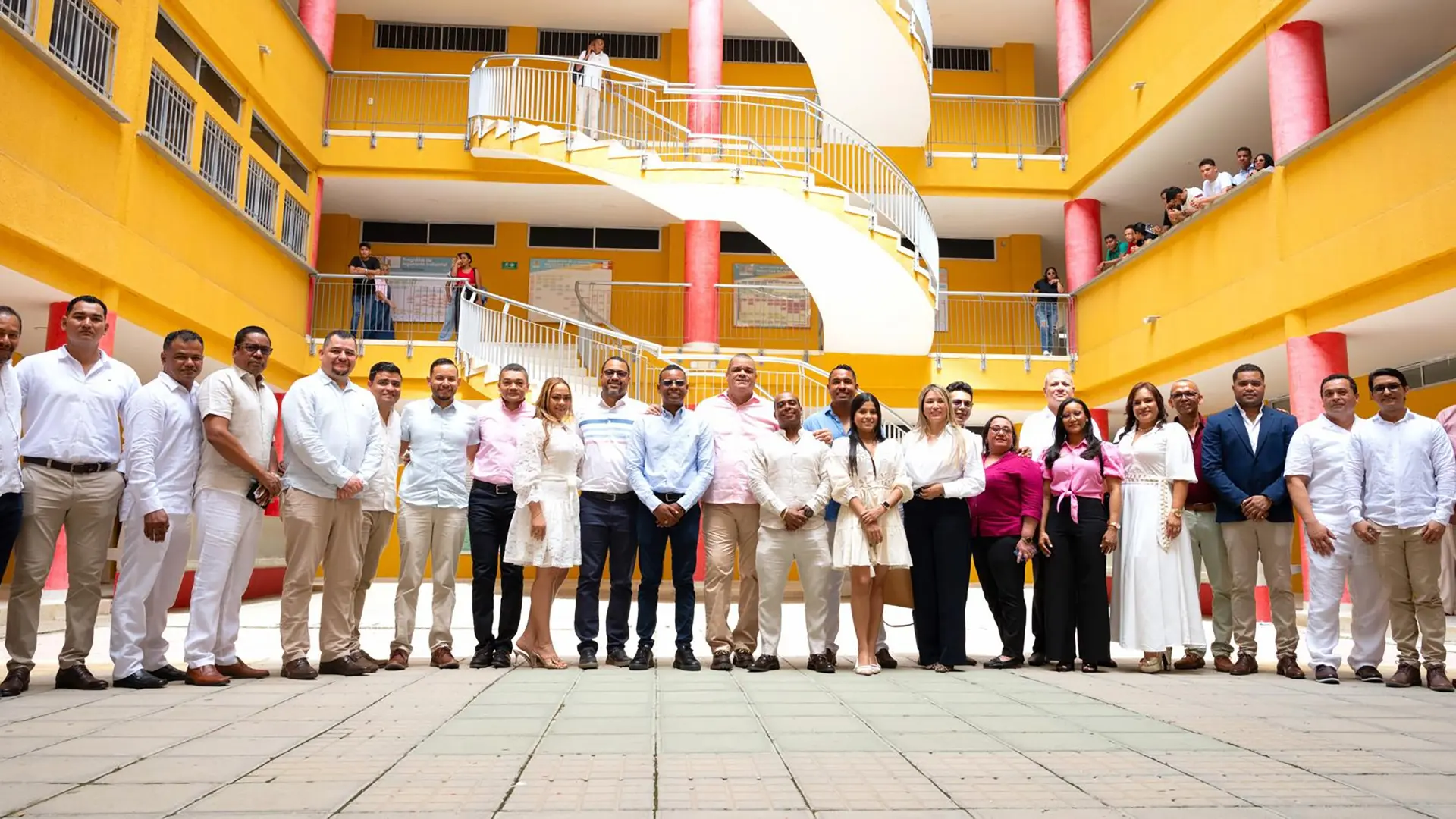 Grupo de personas, entre hombres y mujeres, posan para una foto grupal en el patio central de un edificio institucional con paredes amarillas y columnas rojas. Se trata de un evento formal; la mayoría de los asistentes lleva vestimenta clara y formal. En los niveles superiores del edificio, varias personas observan desde los pasillos.